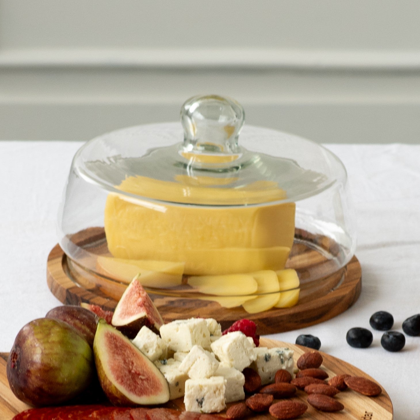 Queijeira com tampa de vidro com queijo e frutas sobre base de madeira