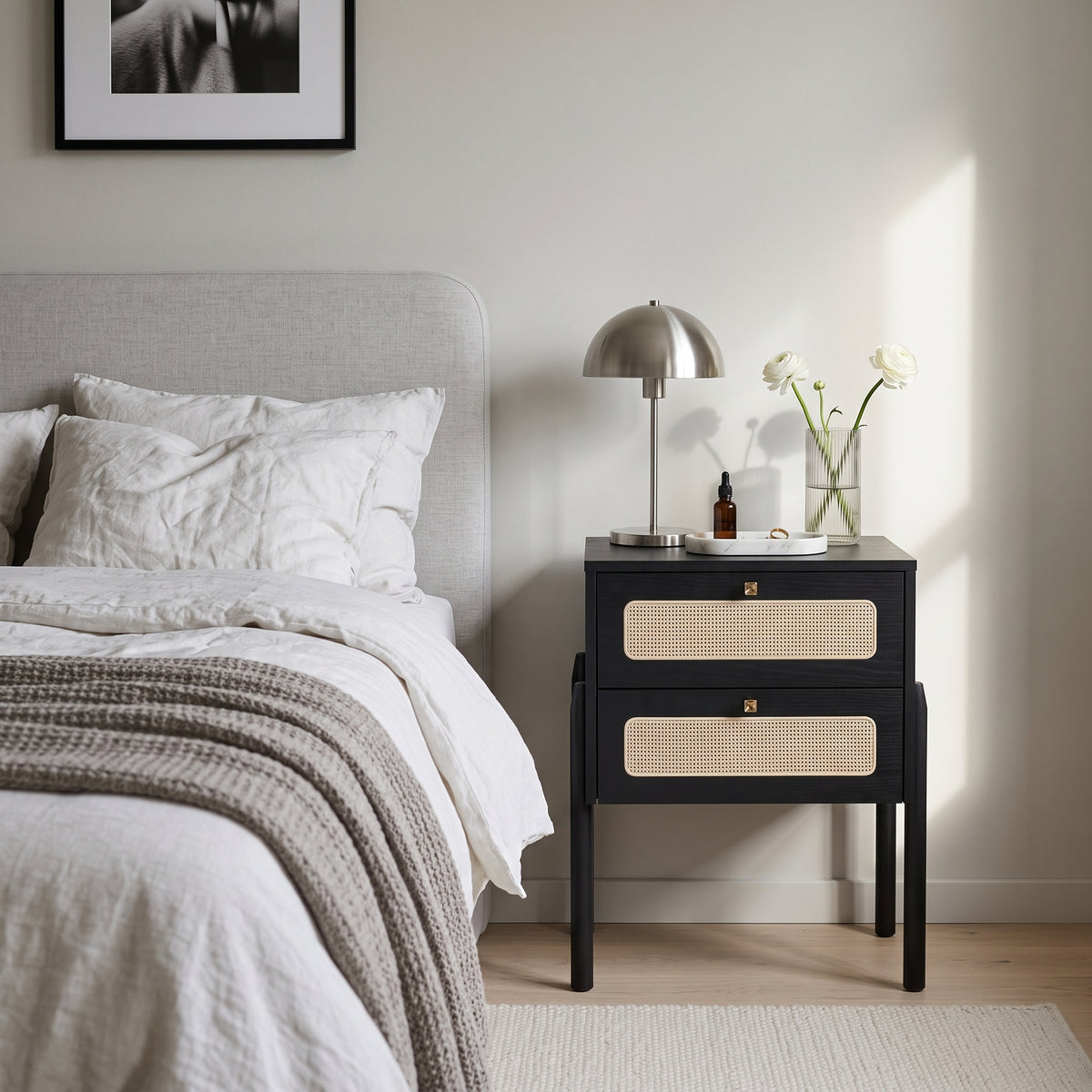 Mesa de cabeceira preta com gavetas de palhinha em quarto com cama, luminária prateada e vaso de flores