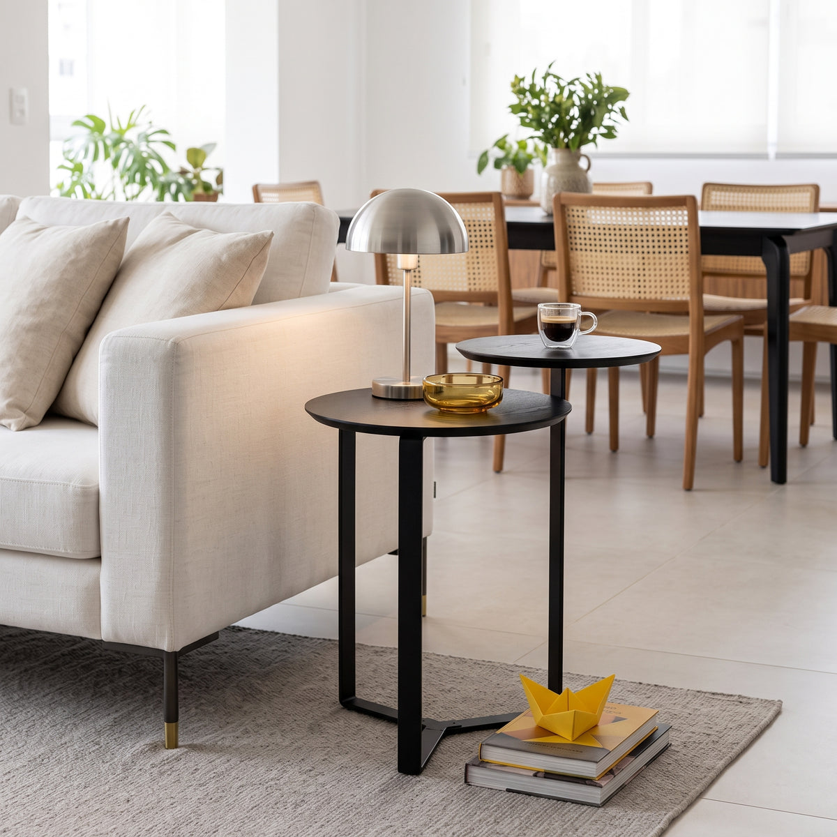 Mesa lateral de dois tampos em sala de estar com sofá claro, luminária prateada e mesa de jantar ao fundo