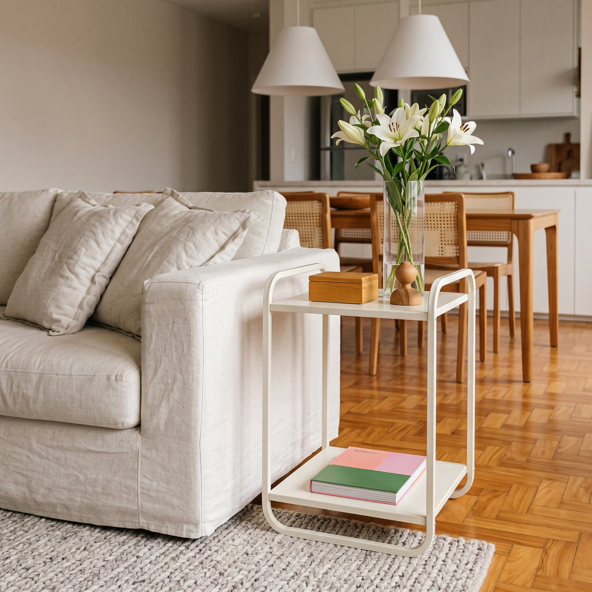 Mesa lateral offwhite ao lado de sofá claro em sala de estar com vaso de lírios e piso de tacos