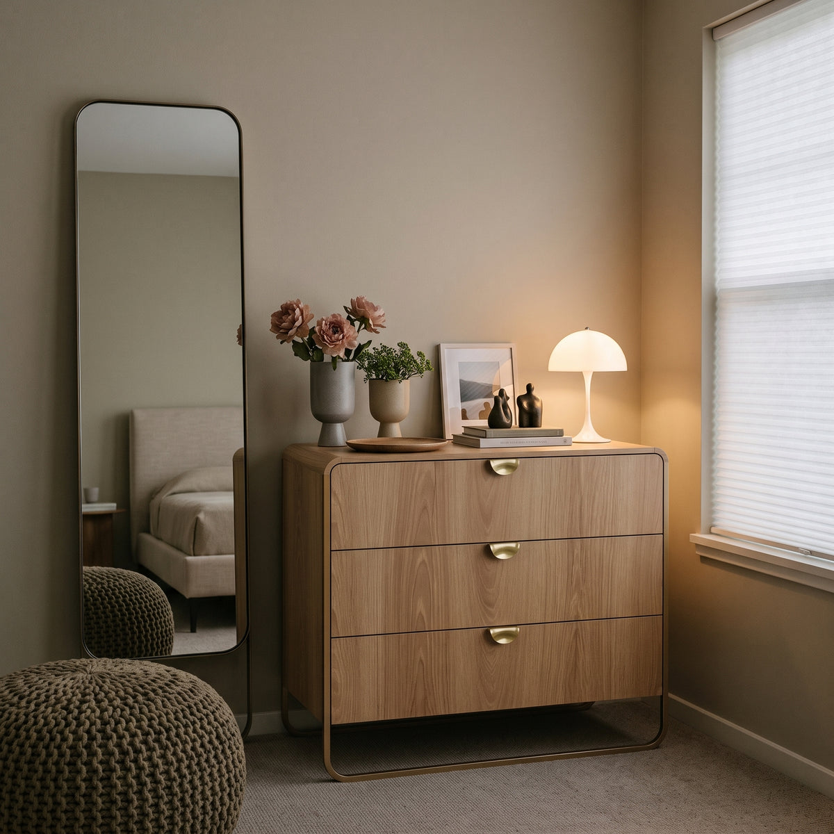 Cômoda amadeirada em quarto com espelho de chão, vaso de flores e luminária de mesa