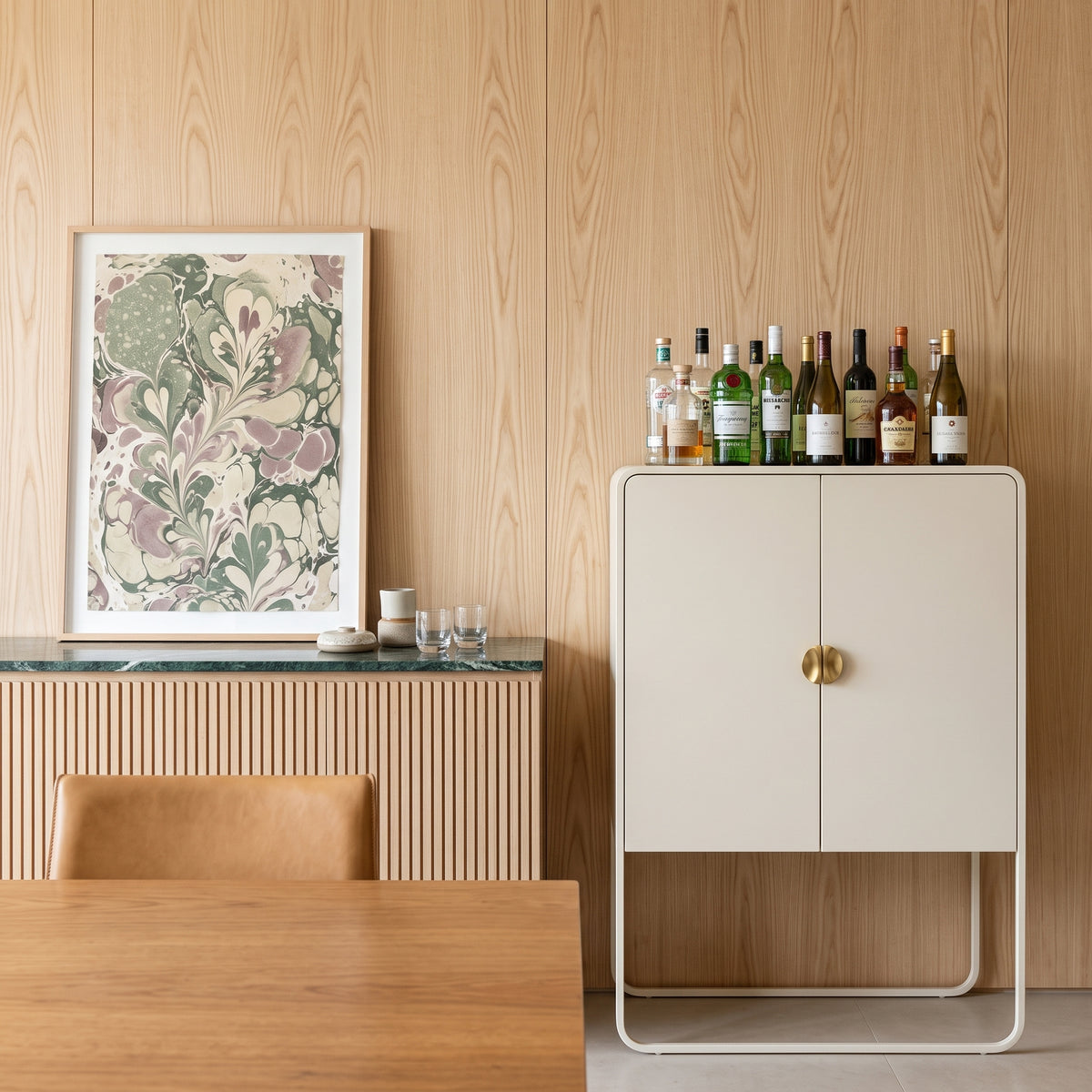 Armário bar offwhite em sala de jantar com garrafas sobre o tampo, quadro floral e painel amadeirado