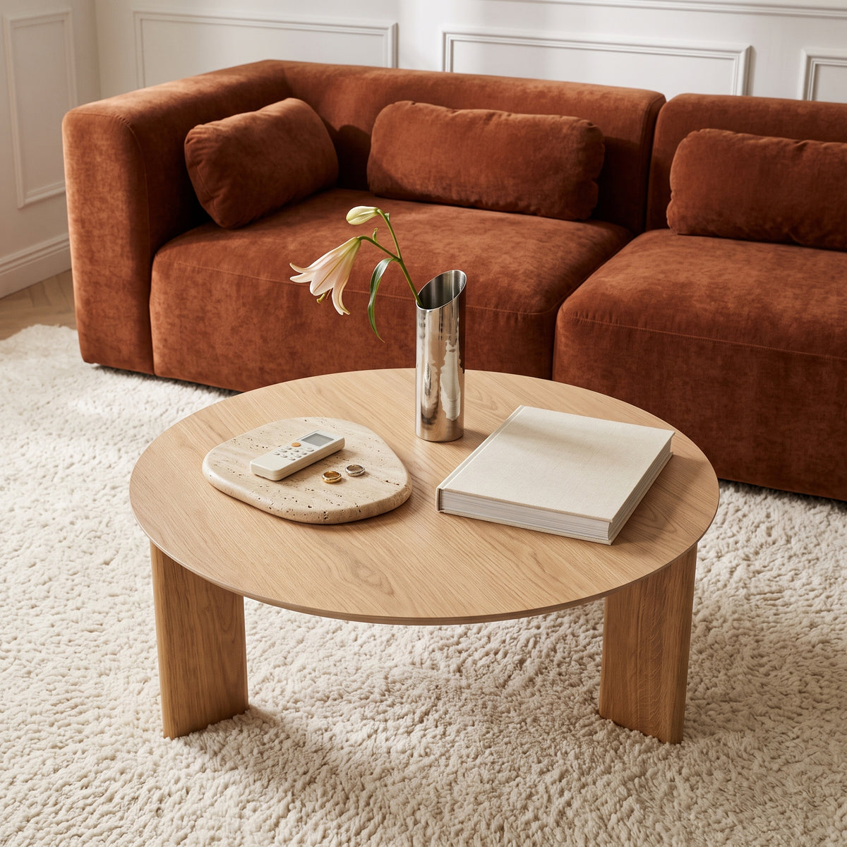 Mesa de centro redonda em sala de estar com sofá de veludo terracota, tapete felpudo claro e vaso com flor