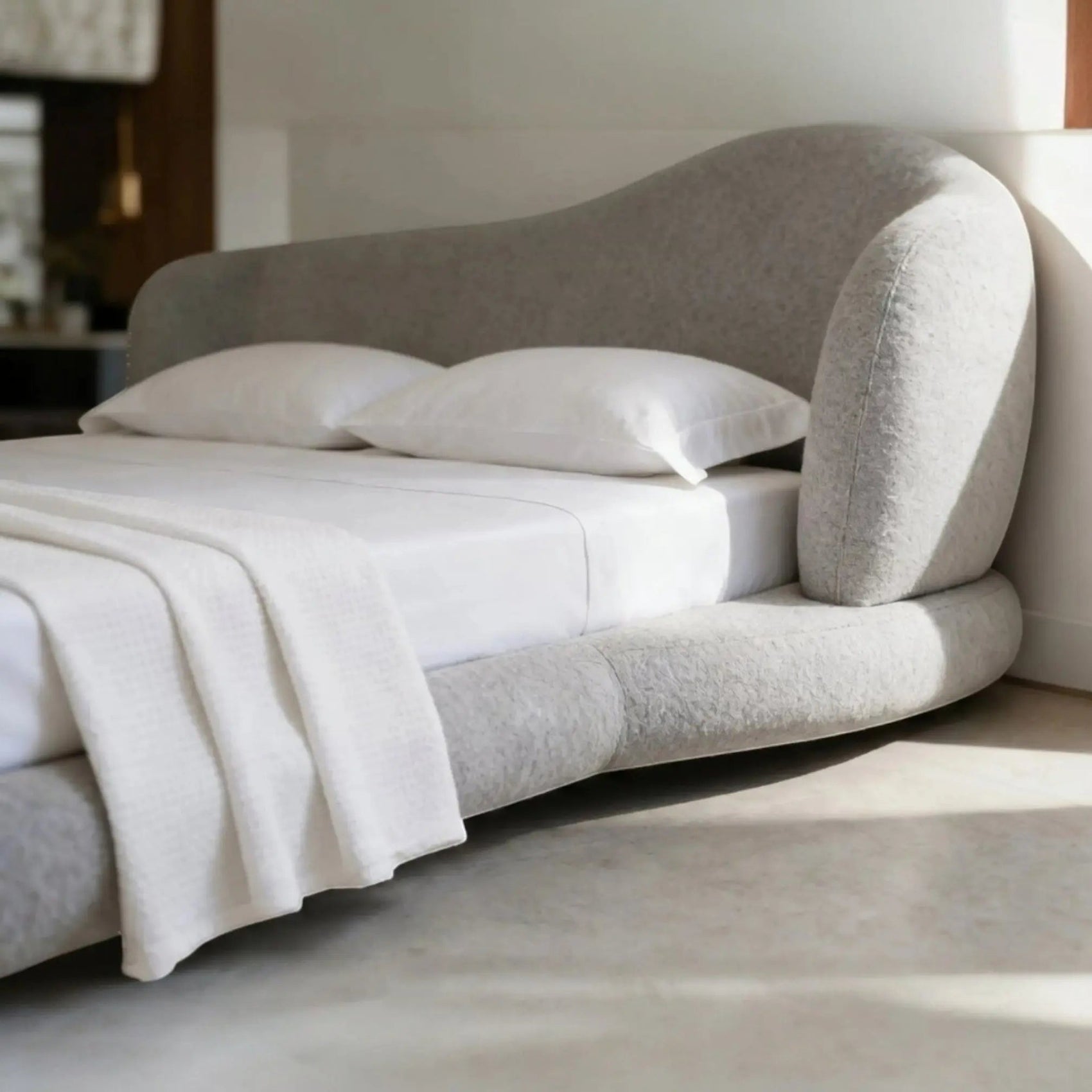 Cama estofada cinza com cabeceira alta curva e almofadas brancas sobre o assento.
