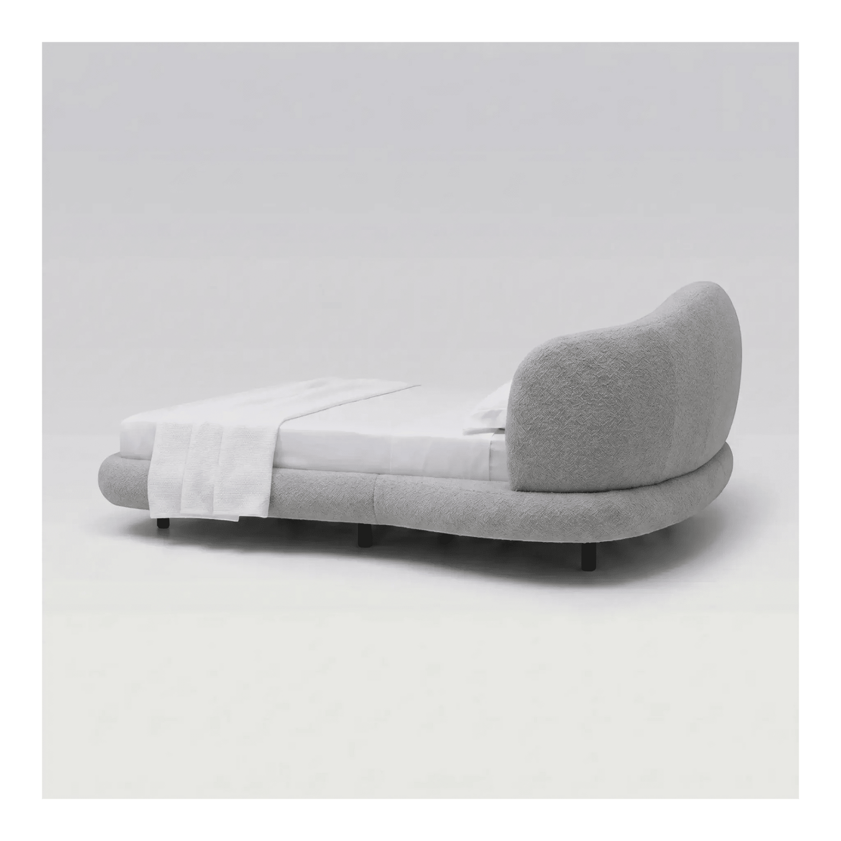 Cama estofada cinza de lado, cabeceira curva e base baixa com travesseiro branco.