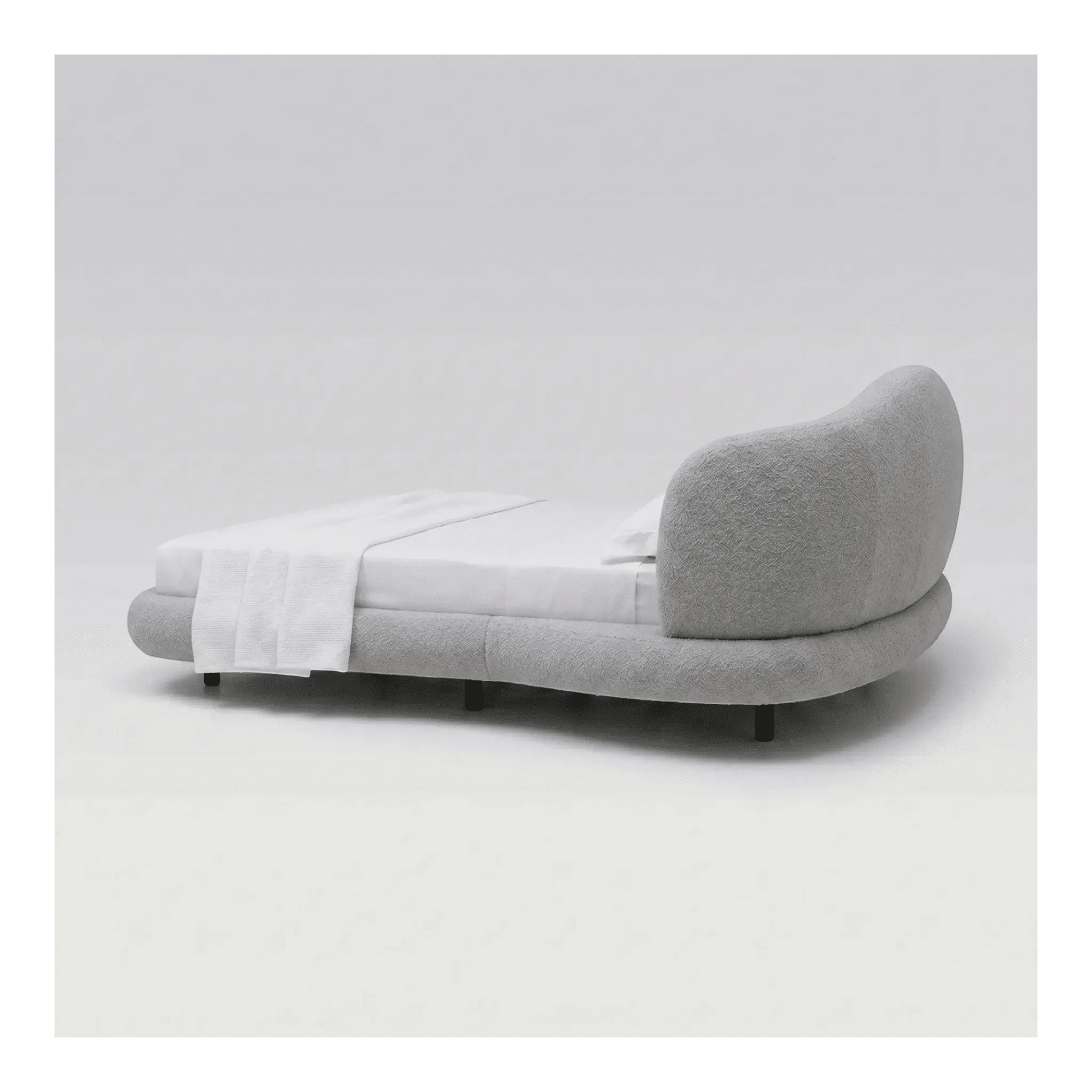 Cama estofada cinza de lado, cabeceira curva e base baixa com travesseiro branco.