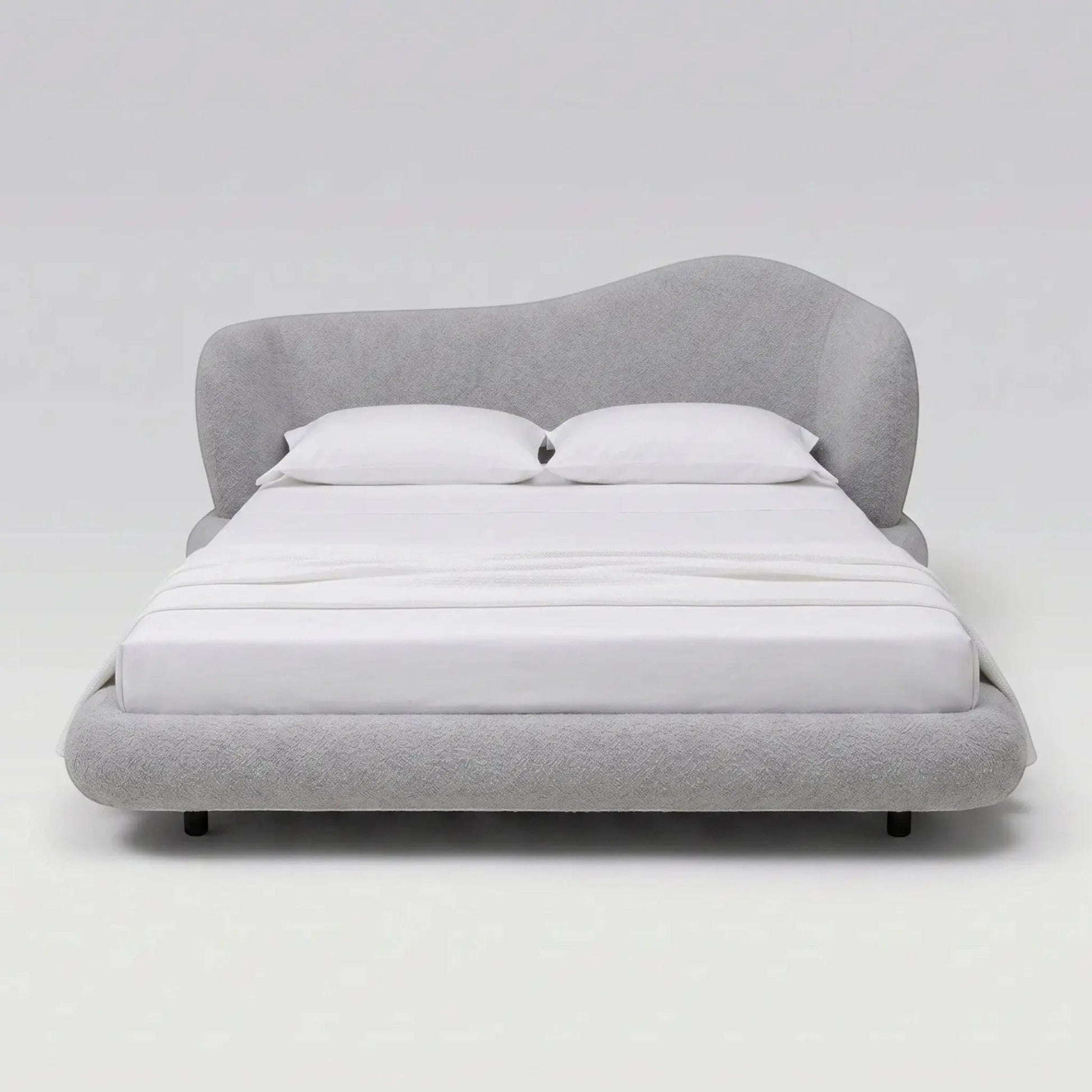 Cama estofada cinza vista de frente, com cabeceira alta arredondada e travesseiros brancos.