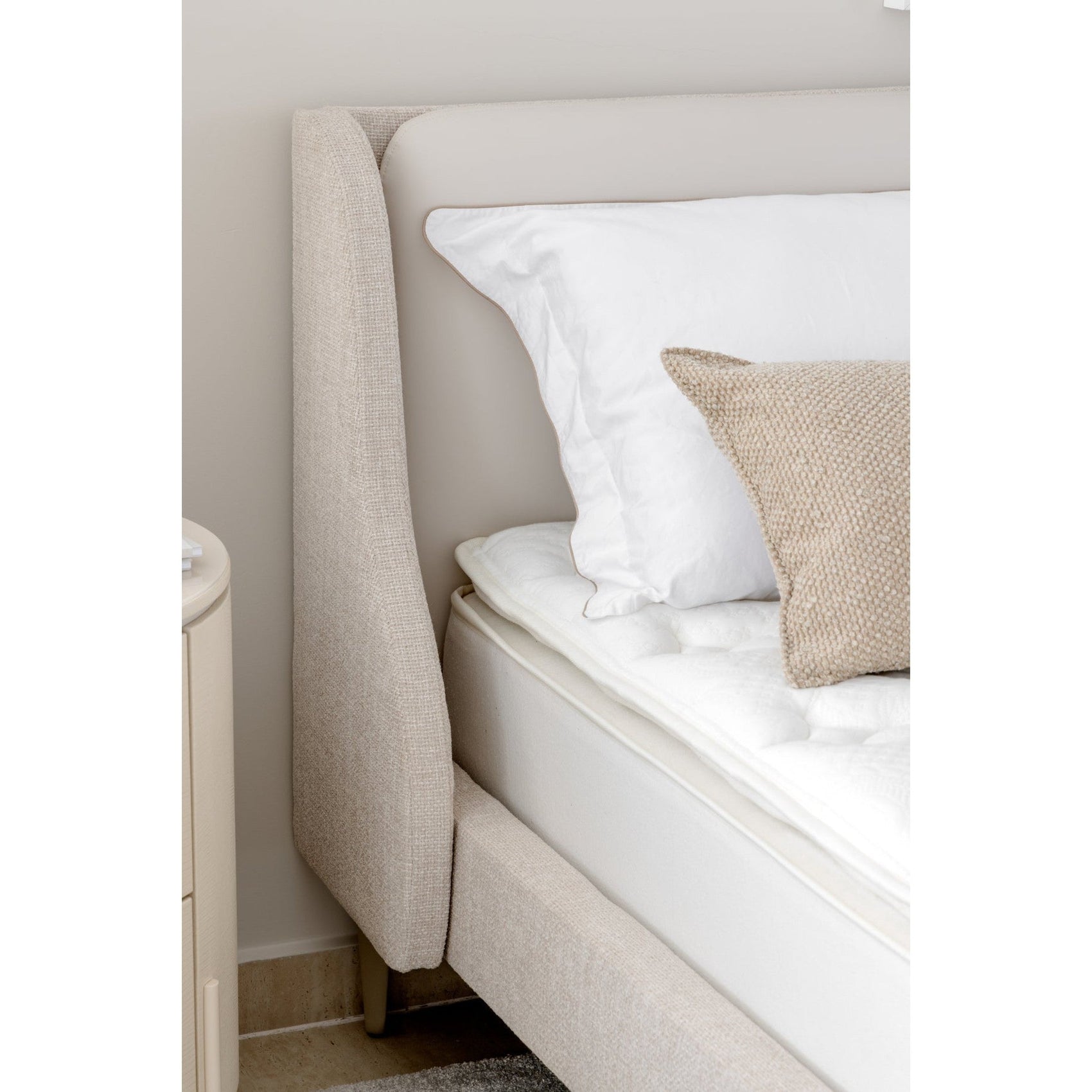 Cama estofada bege com lateral alta e travesseiros brancos, almofada bege texturizada.