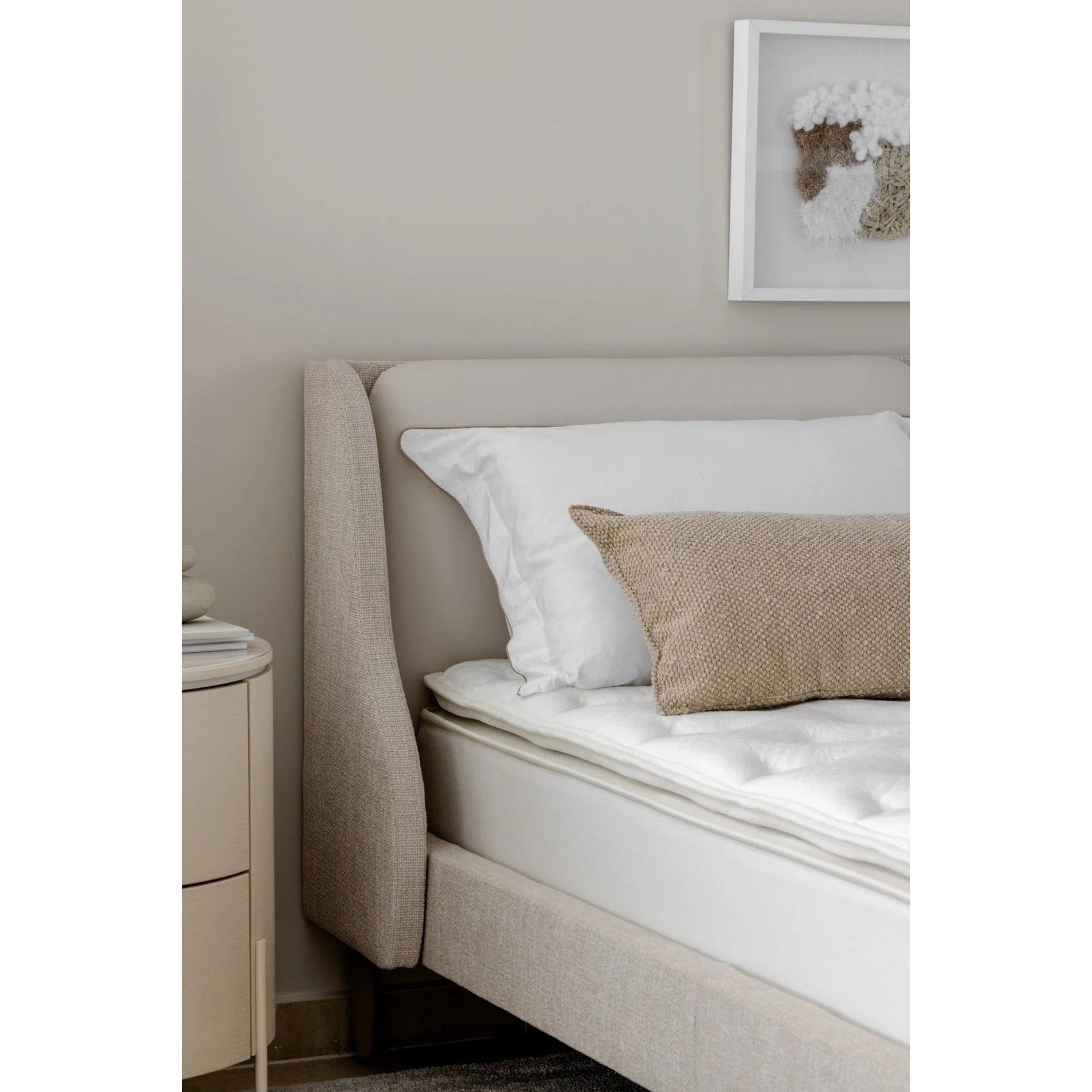 Cama estofada lateral bege, com colchão e almofadas brancas e capa bege texturizada.