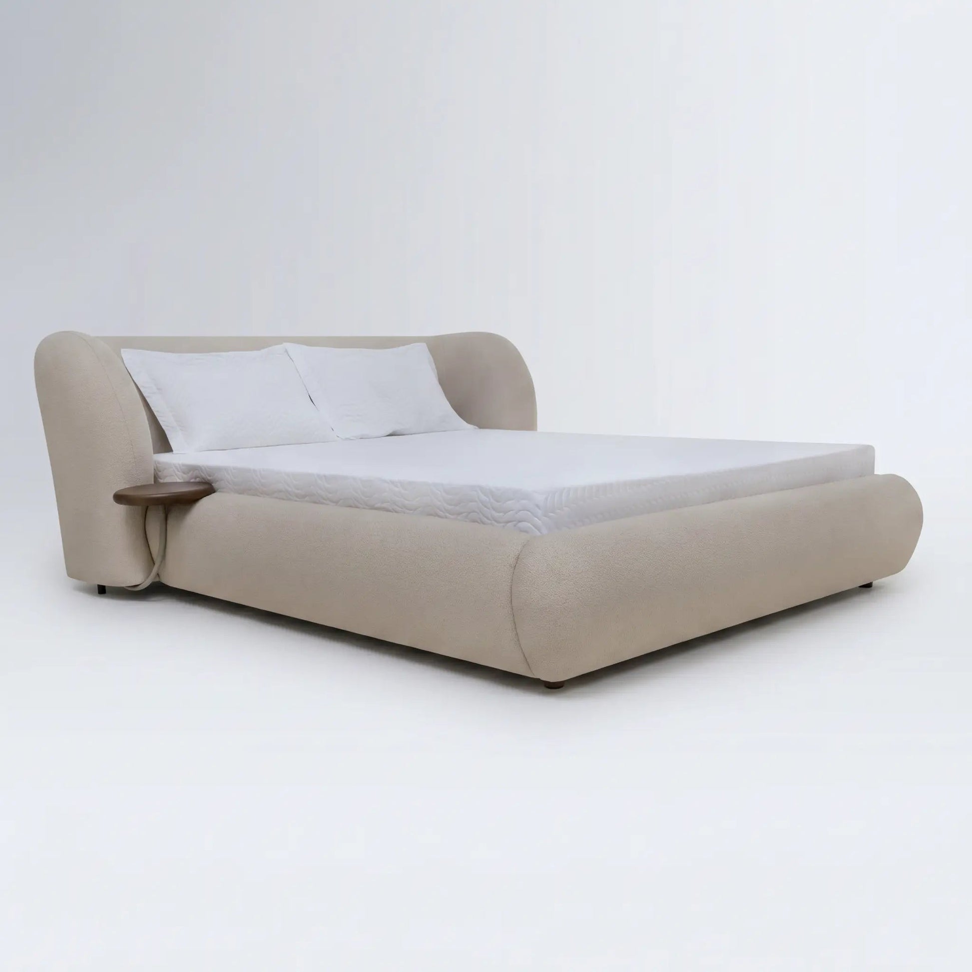Cama estofada bege com cabeceira alta curva, almofadas brancas e mesa lateral