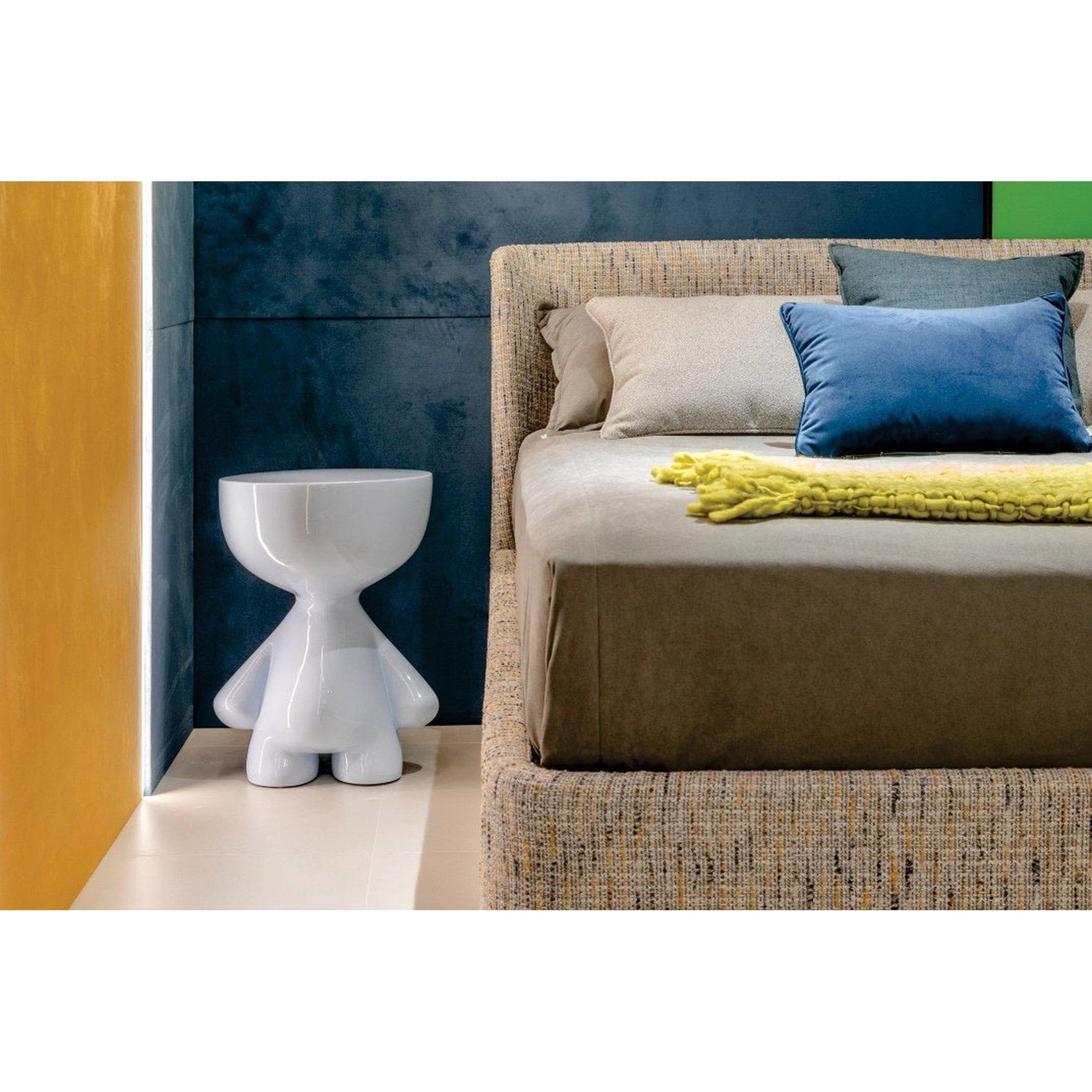 Cama estofada cinza com roupa branca, travesseiro azul e manta amarela sobre o leito