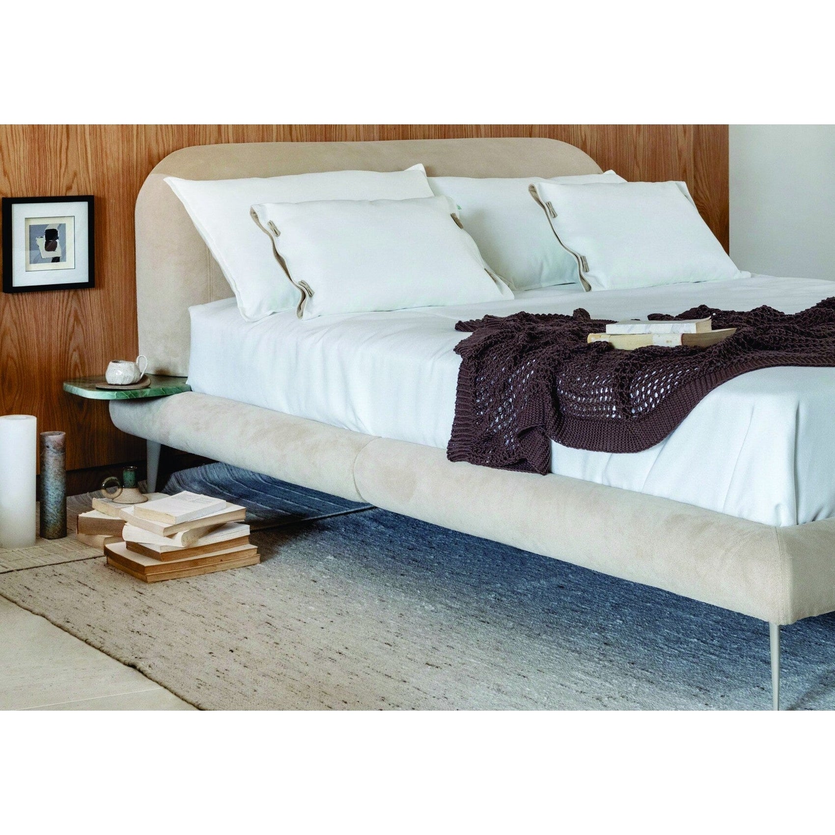 Cama estofada bege com lençóis e travesseiros brancos, manta escura dobrada sobre a cama